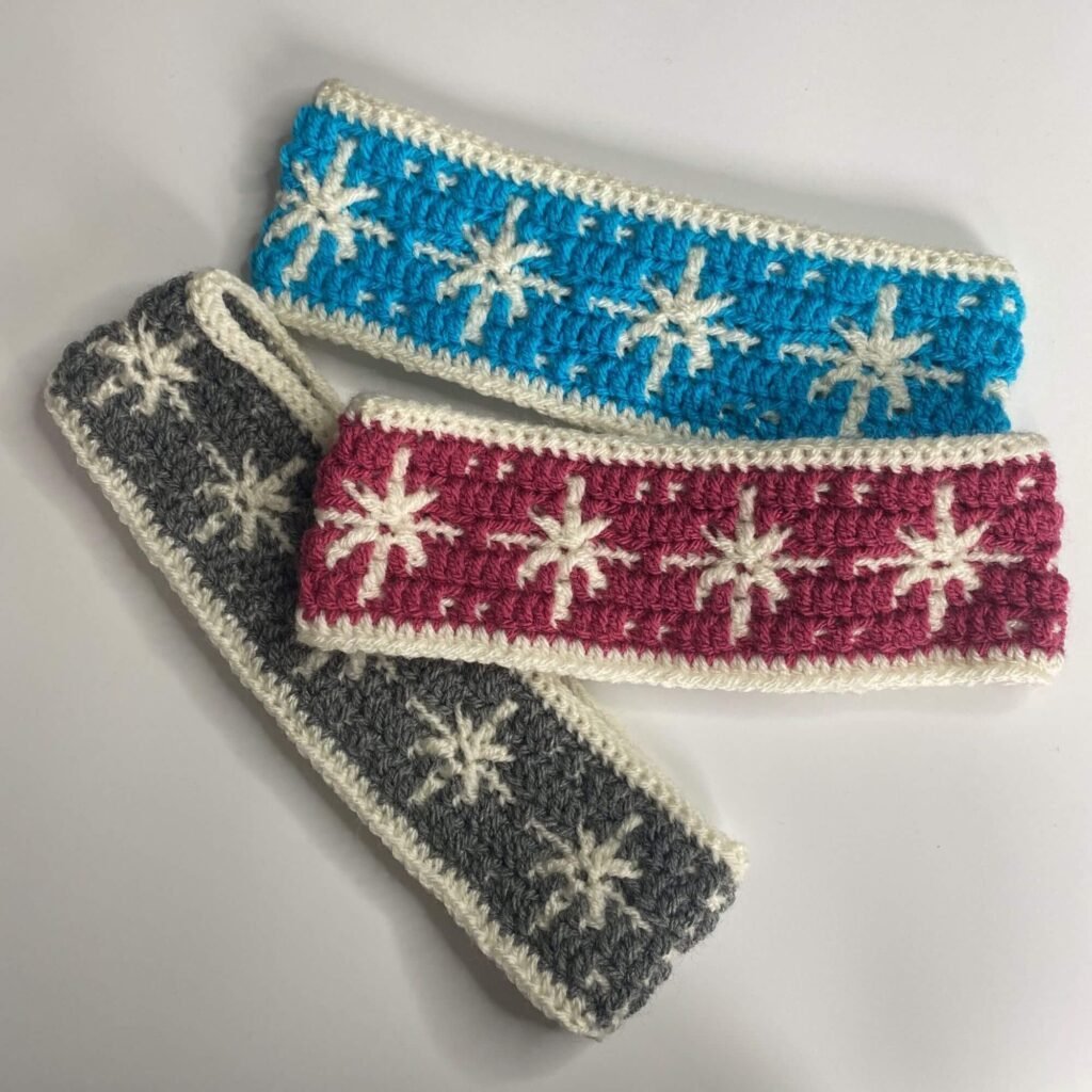 Three crocheted headbands with snowflakes on them in blue, pink and grey