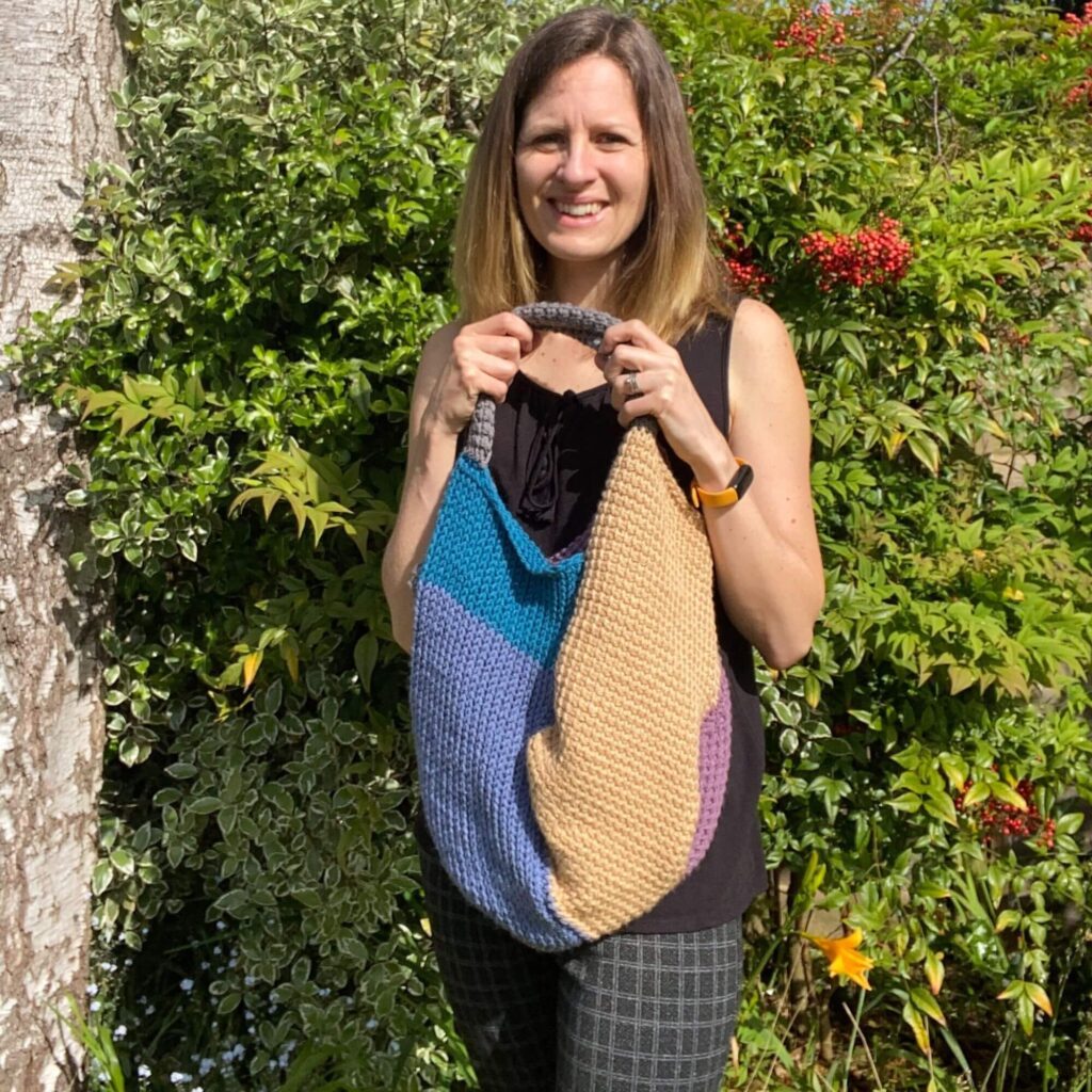 A lady holds up a scrap crochet bag in different colour blocks made using Tunisian Crochet