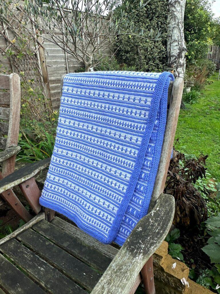 A blue and white crochet blanket hangs over a wooden chair in a garden
