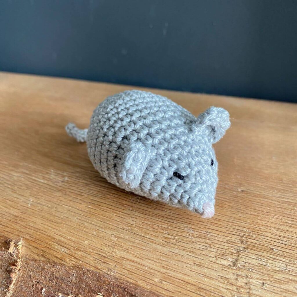 A grey amigurumi mouse sat on a wooden shelf
