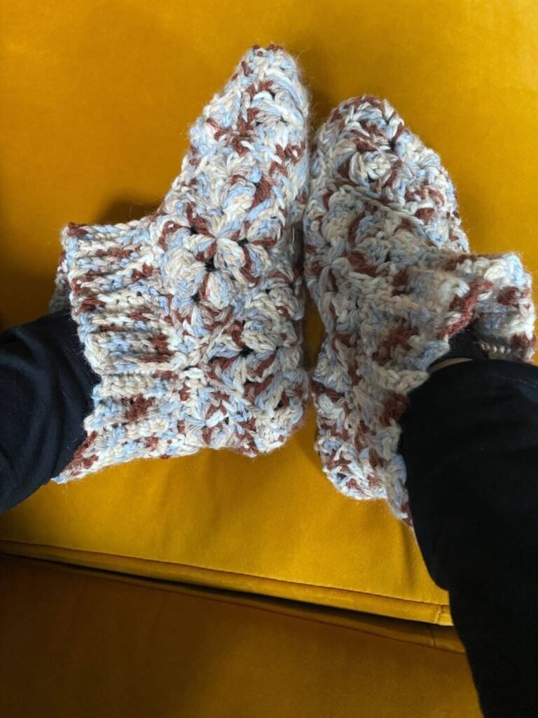 A pair of crochet slippers in brown and blue variegated yarn worn by someone with black leggings against a yellow sofa background