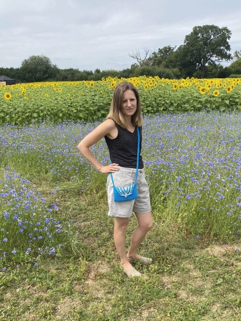 A lady in a field is wearing shorts and a vest and a crochet blue and beige cross body handbag