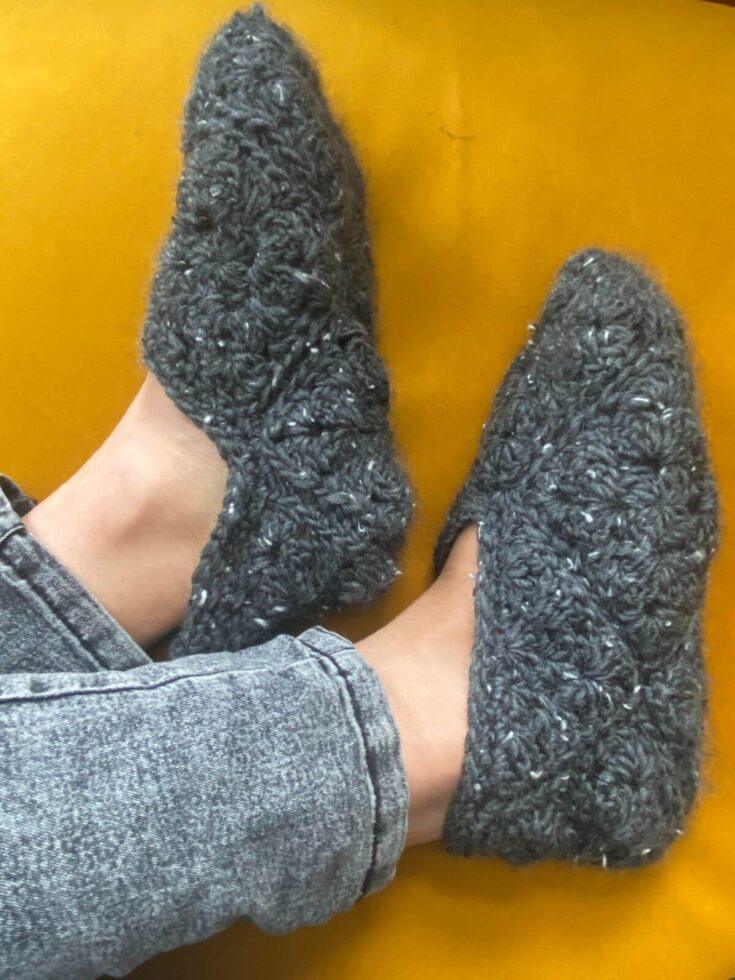 A pair of crochet slippers shown side on worn on feet on a yellow sofa