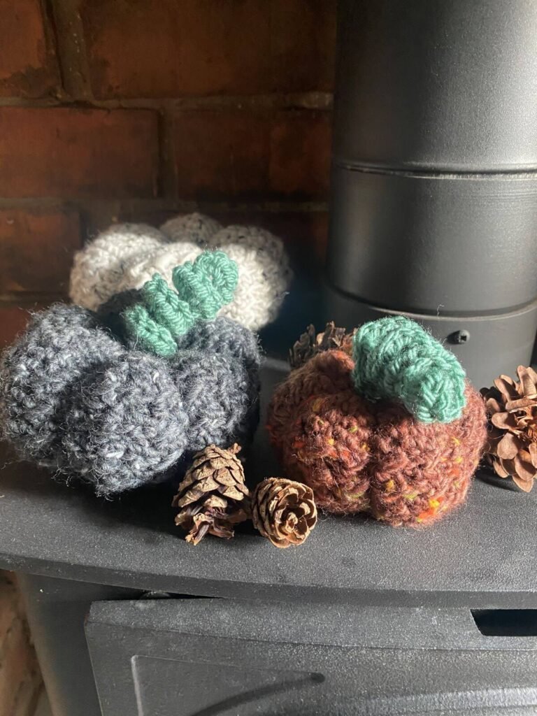 3 crochet pumpkins on a fireplace surrounded by pinecones. The pumpkins are red, grey and white with green curly stems