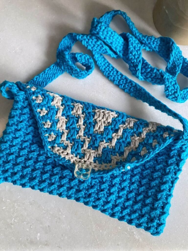 A blue and cream crochet bag lying on a white surface. The bag has a mosaic crochet detail on the bag flap and a blue long strap