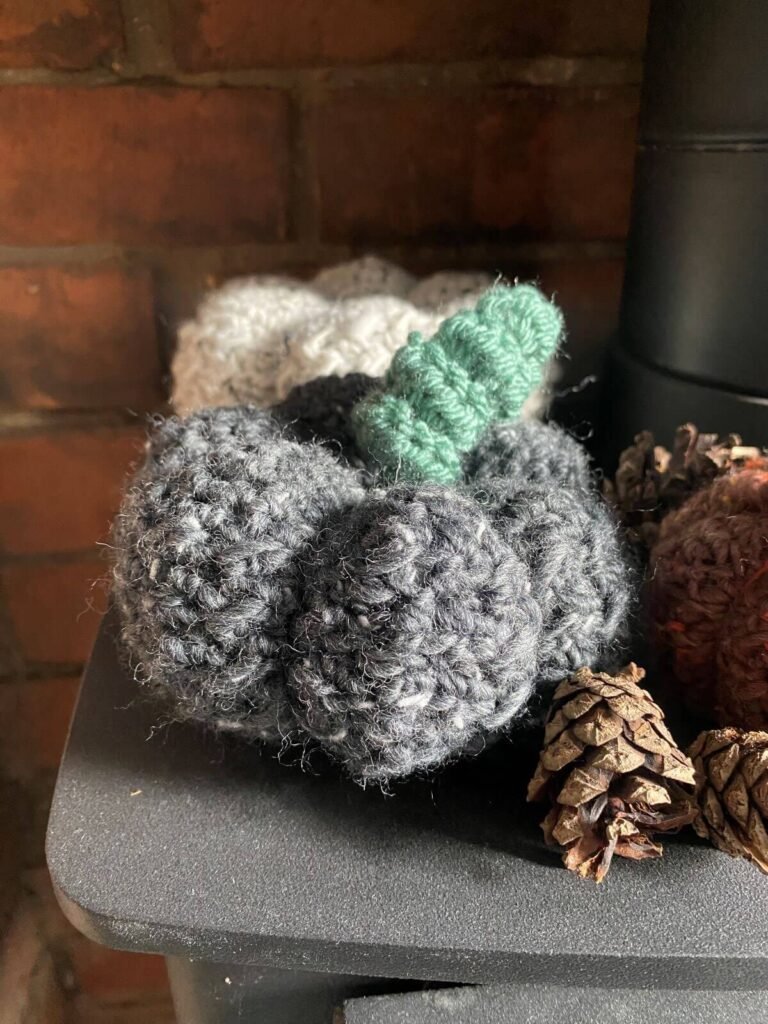 A large grey crochet pumpkin sits on a hearth next to some pine cones and other crochet pumpkins