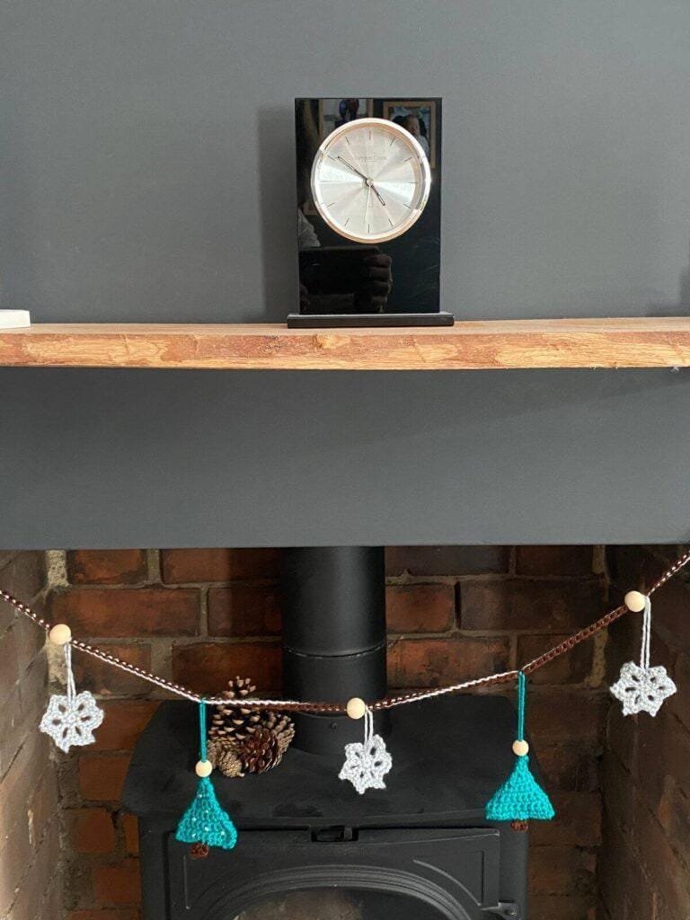 A crochet christmas garland hangs on a fireplace with a wooden mantelpiece, a black clock and a log burner