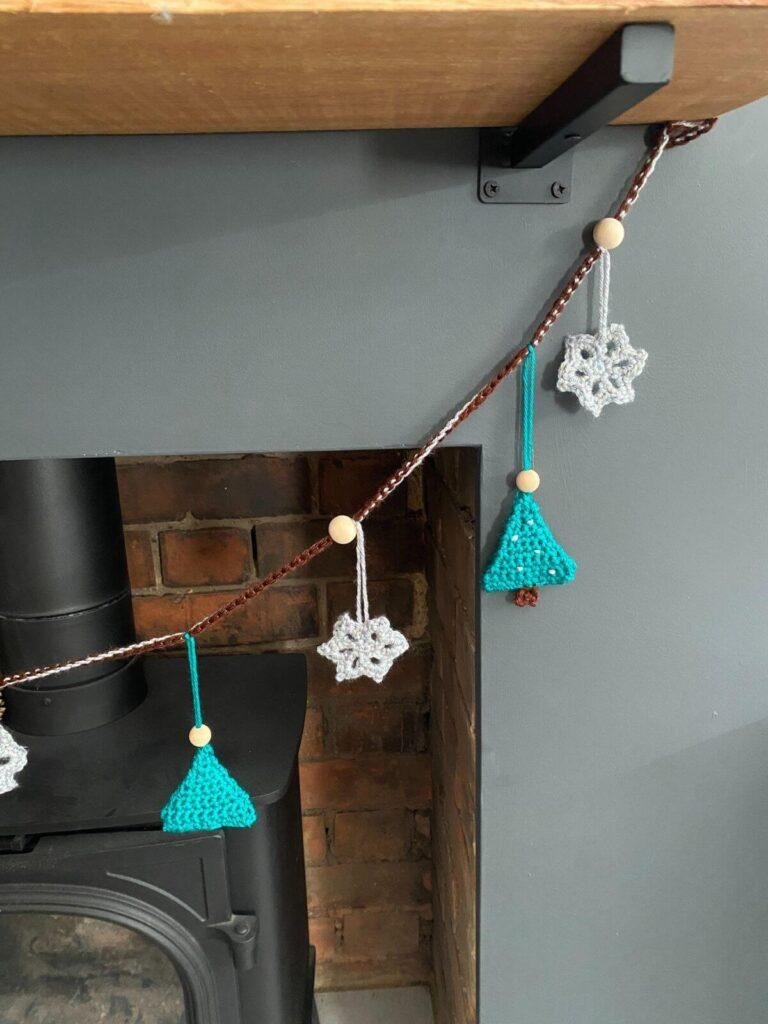 A close up of a fireplace showing half of the log burner, a wooden mantel and a crochet Christmas garland of trees and snowflakes hanging from it
