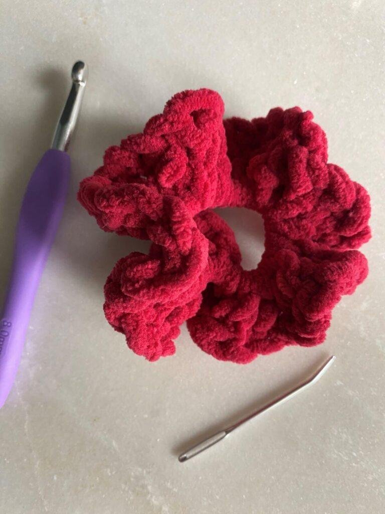 A red crochet scrunchie on a grey surface next to a crochet hook and needle