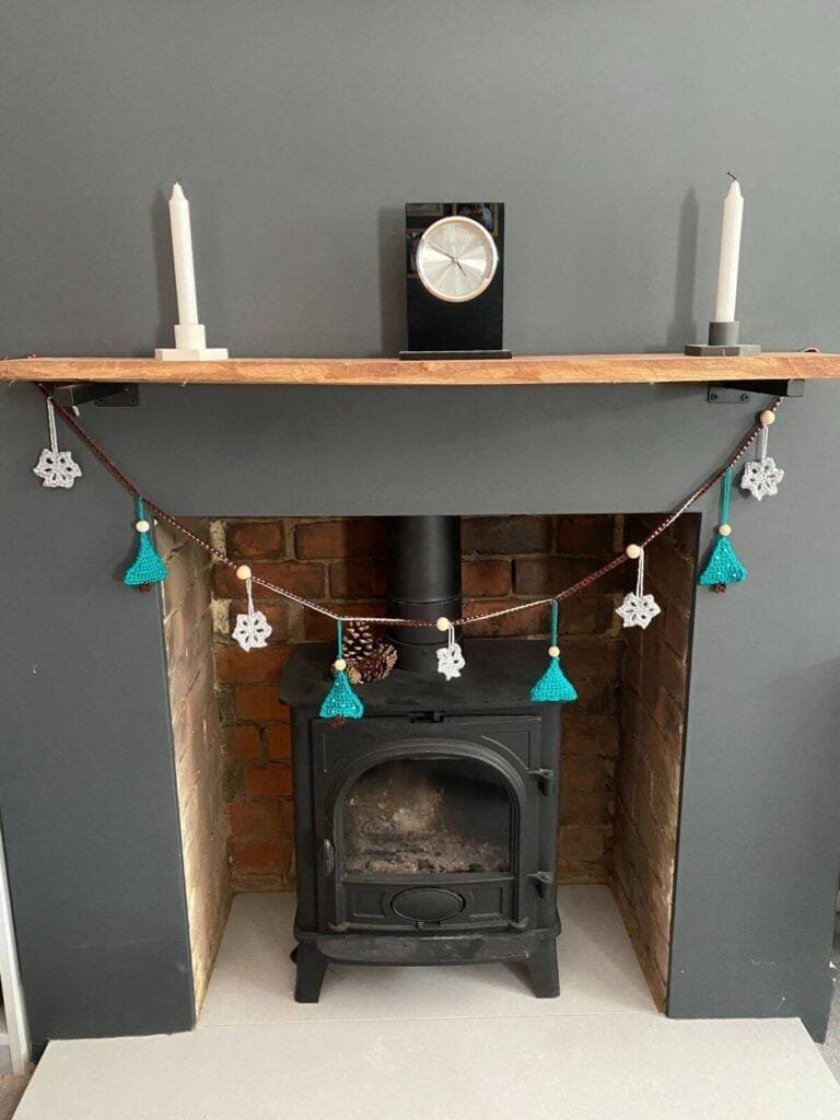 A fireplace with candles and a clock on a wooden mantelpiece and a crochet garland hanging underneath featuring Christmas trees and snowflakes