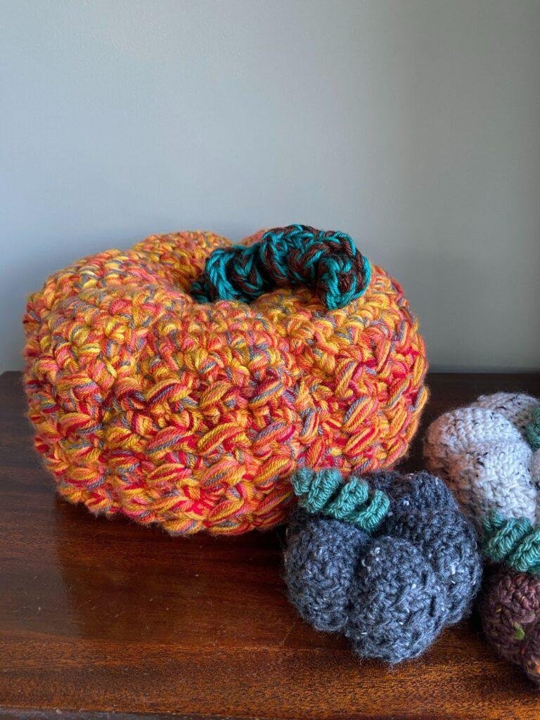 A big orange crochet pumpkin on a wooden table with a small grey and white pumpkin next to it