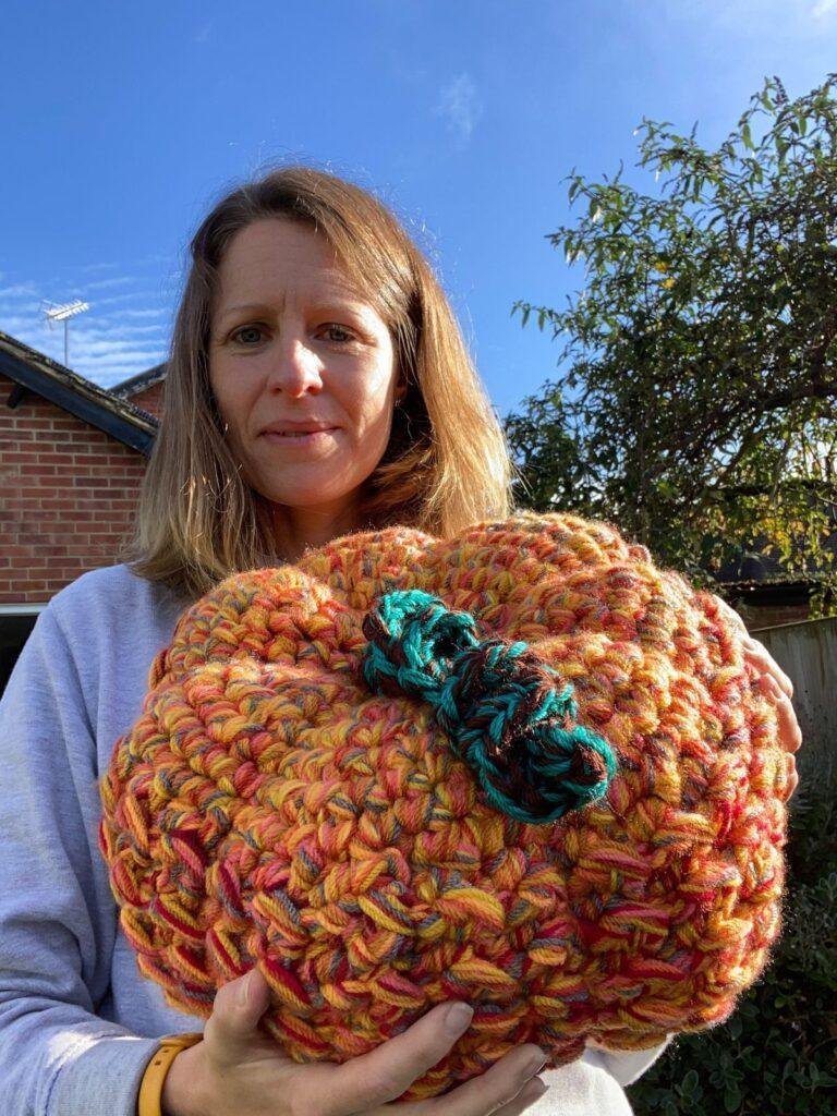 A lady stands in a garden holding a giant orange crochet pumpkin