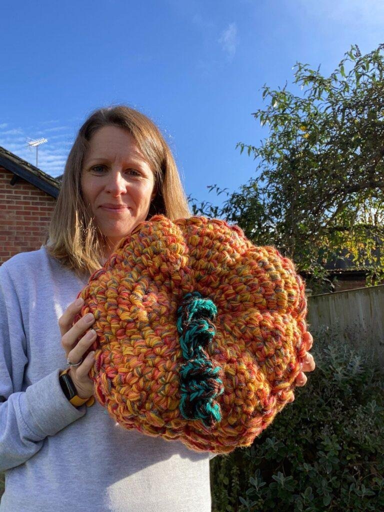 A lady stands in a garden holding a large crochet pumpkin she has made