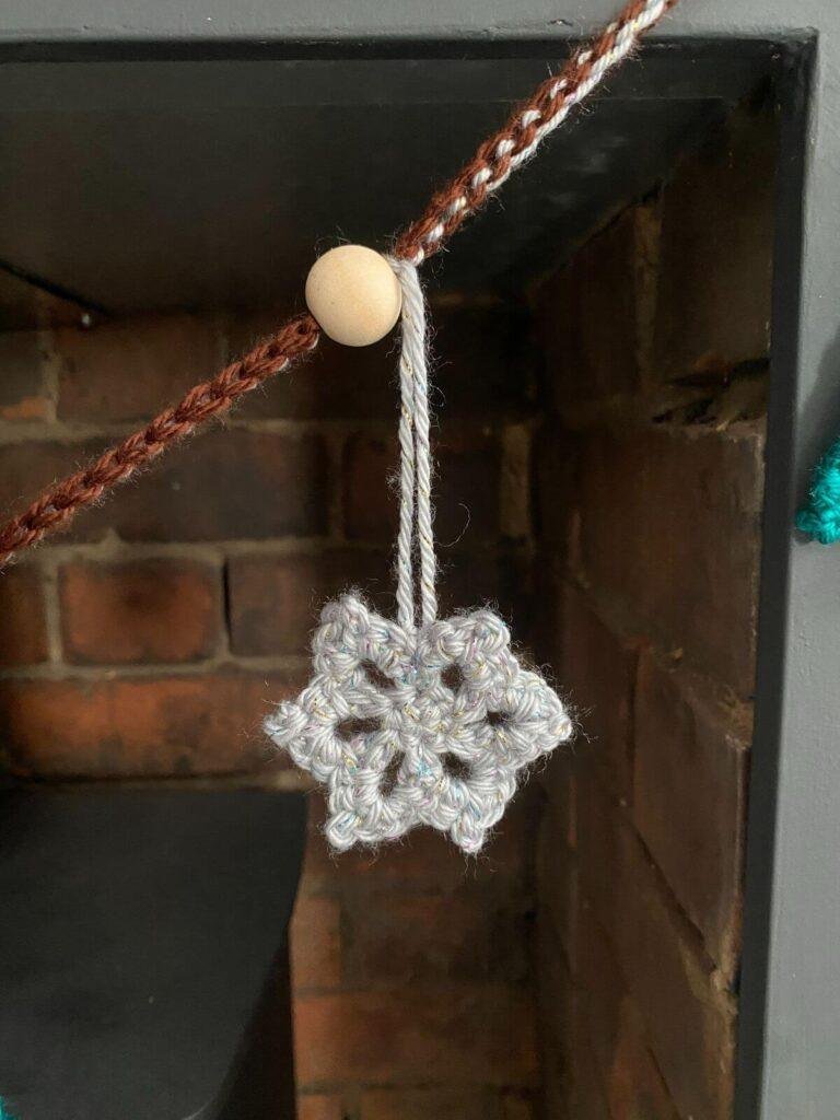 A crochet snowflake in silver hangs from a garland against a brick background