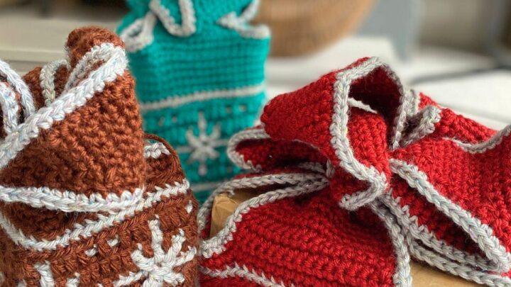 3 crochet gift boxes with snowflake motifs in red, brown and green sit on the floor