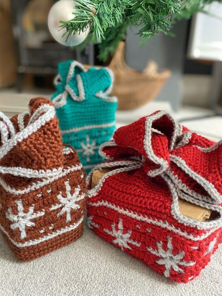 3 crochet gift boxes with snowflake motifs in red, brown and green sit on the floor