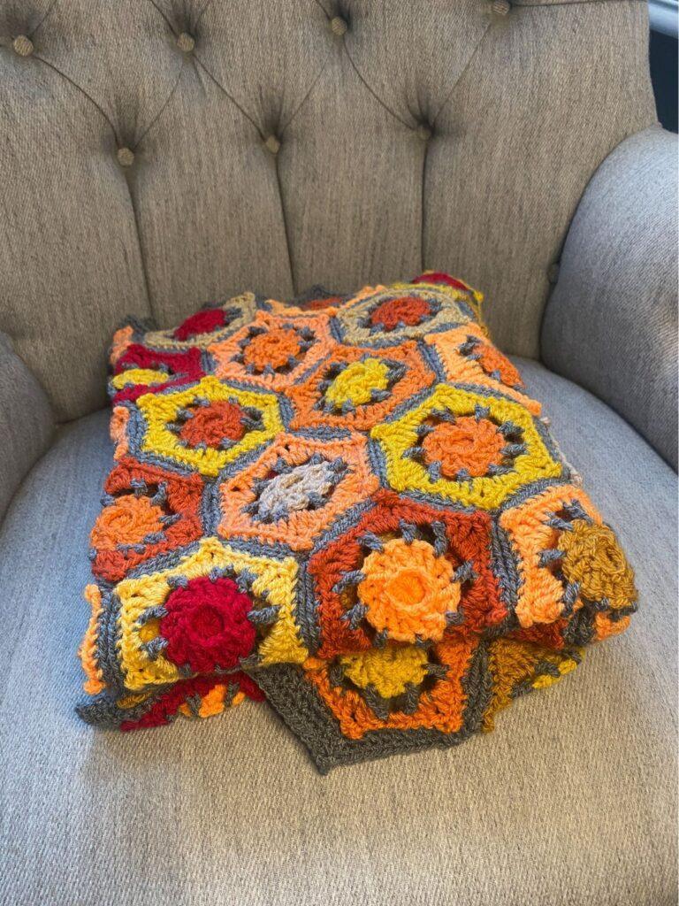 A folded crochet blanket in red, orange and grey is sat on an armchair