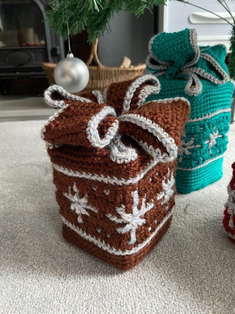 A brown crochet gift box with silver snowflakes sits on a carpet in front of other crochet boxes