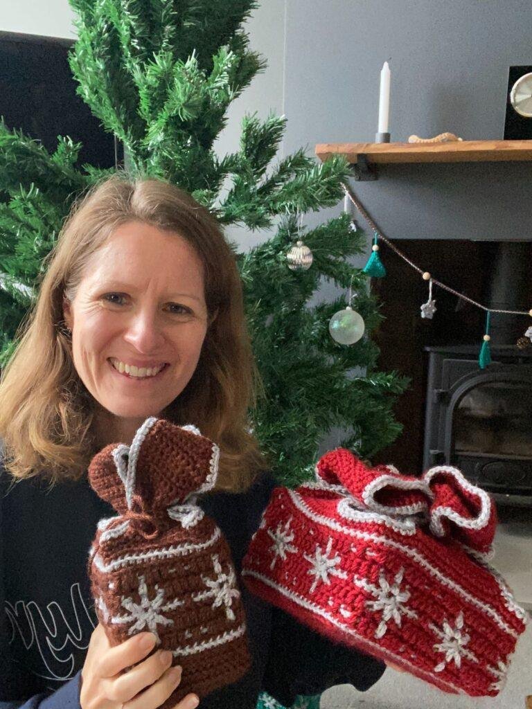 A lady is in front of a Christmas tree holding a red and a brown crochet gift box with silver snowflakes on