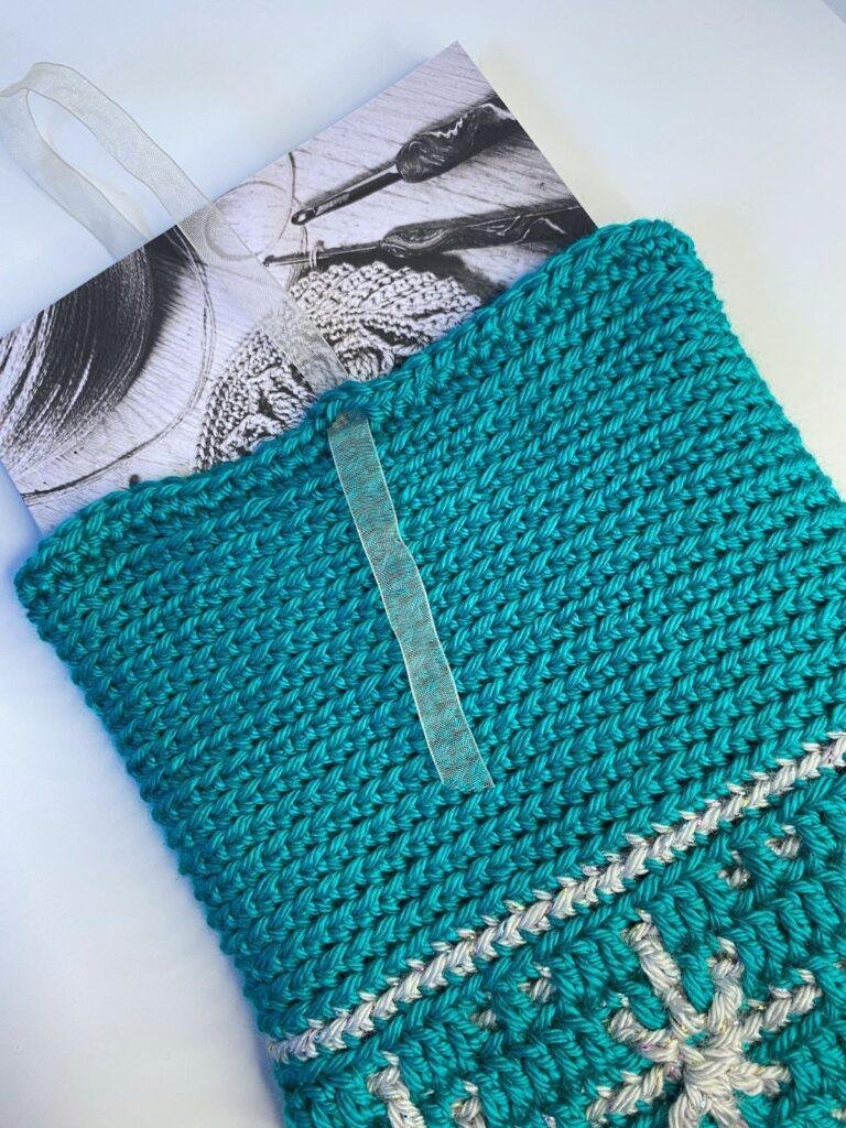 A festive crochet book cover with a snowflake on it in silver, has a book poking out of the top and a ribbon