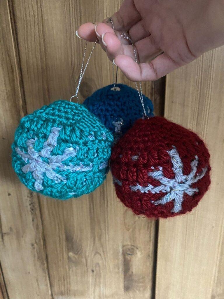 3 crochet baubles in red, green and blue with a snowflake on them are held in a hand in front of a wooden door