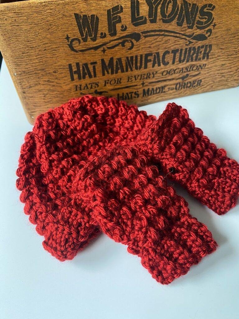 A pair of textured red crochet mittens and a red crochet hat sit next to a wooden box with hat manufacturer written on it