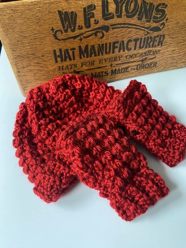 A pair of textured red crochet mittens and a red crochet hat sit next to a wooden box with hat manufacturer written on it