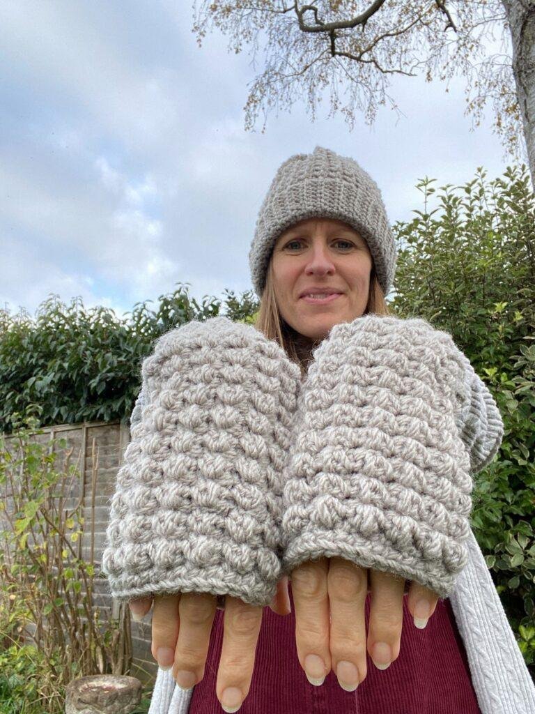 A lady standing outside is wearing crochet hat and mittens in grey holding out the mittens to the camera