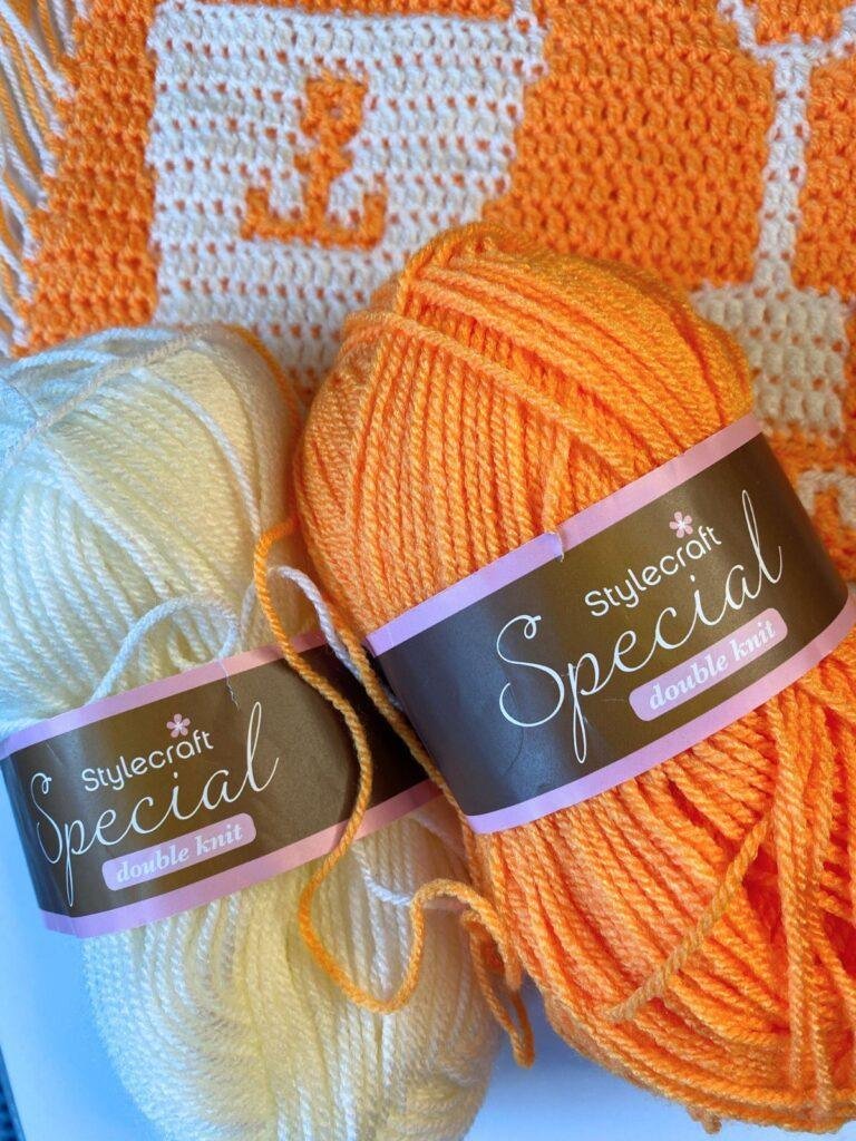 2 balls of yarn sit on top of a crochet strip showing a bucket in orange and white