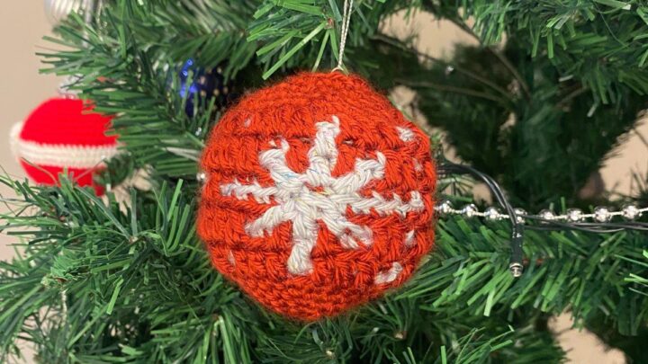 A red crochet bauble with a silver snowflake hangs on a decorated Christmas tree