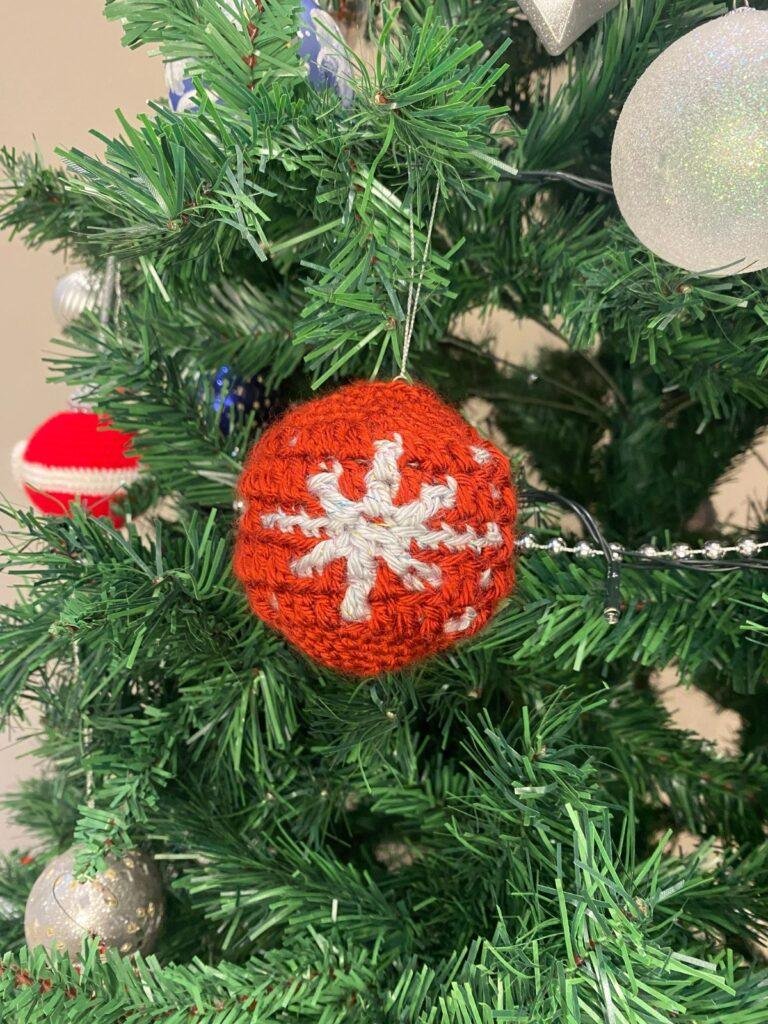 A red crochet bauble with a silver snowflake hangs on a decorated Christmas tree