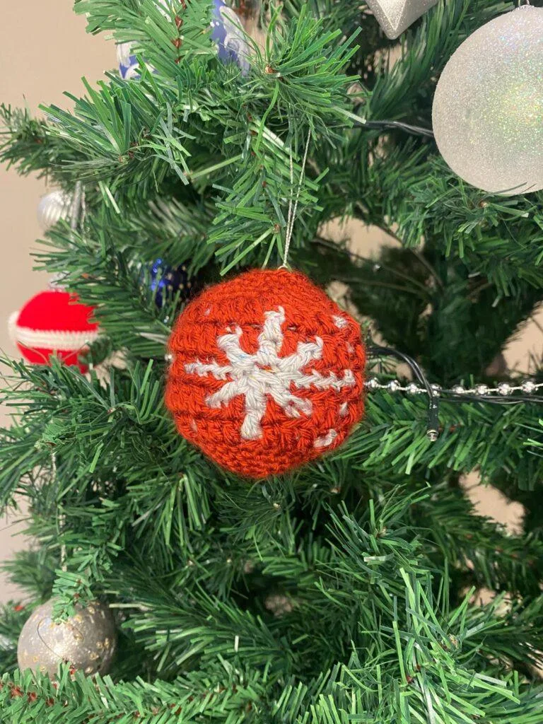 A red crochet bauble with a silver snowflake hangs on a decorated Christmas tree