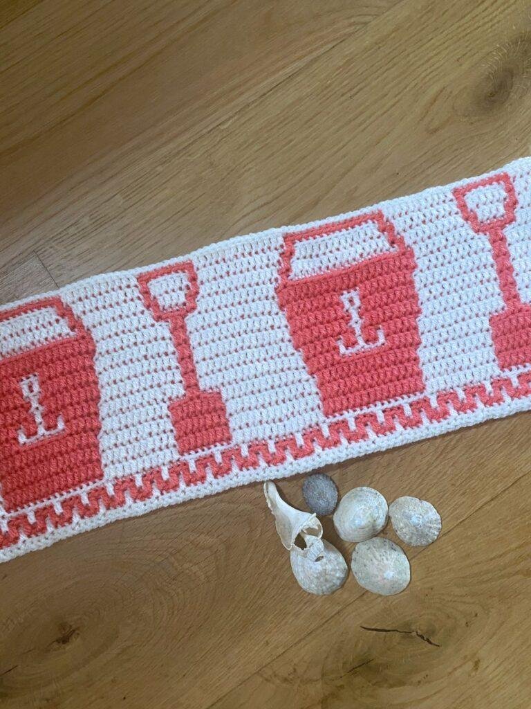 A pink and white crochet strip with a bucket and spade motif