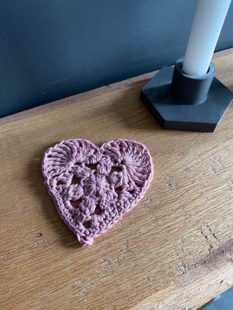 A pink crochet love heart on a wooden shelf with a black candle holder behind