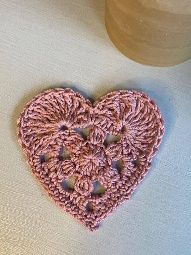A pink crochet heart motif on a white background with a beige jar on the edge