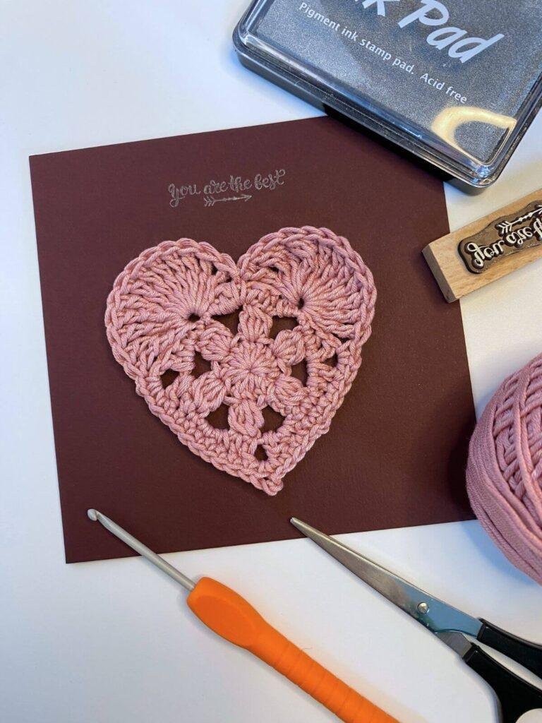 A crochet heart on a Valentine's Day card with crochet tools around the edge
