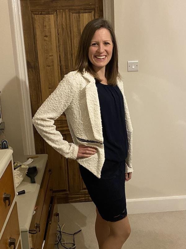 A woman stands in a bedroom with a blue dress and white jacket with her hand on her waist, smiling