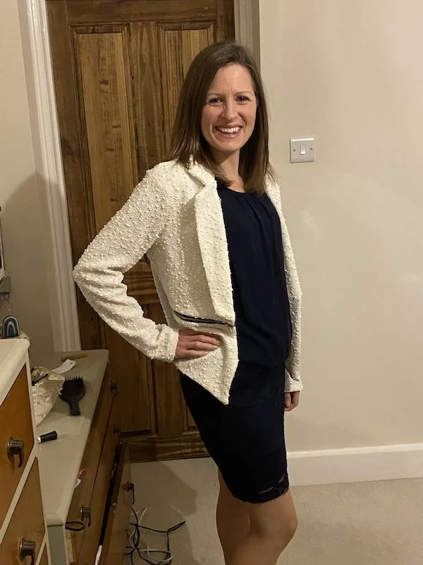 A woman stands in a bedroom with a blue dress and white jacket with her hand on her waist, smiling