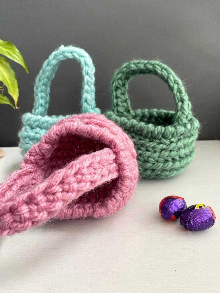 3 chunky crochet baskets on a white surface with 2 mini easter eggs next to them. One basket is lying down