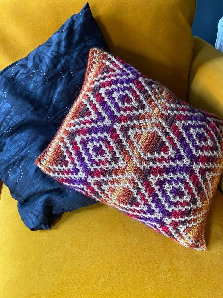 A bright patterned crochet pillow, sits in front of a black pillow, both on a yellow couch