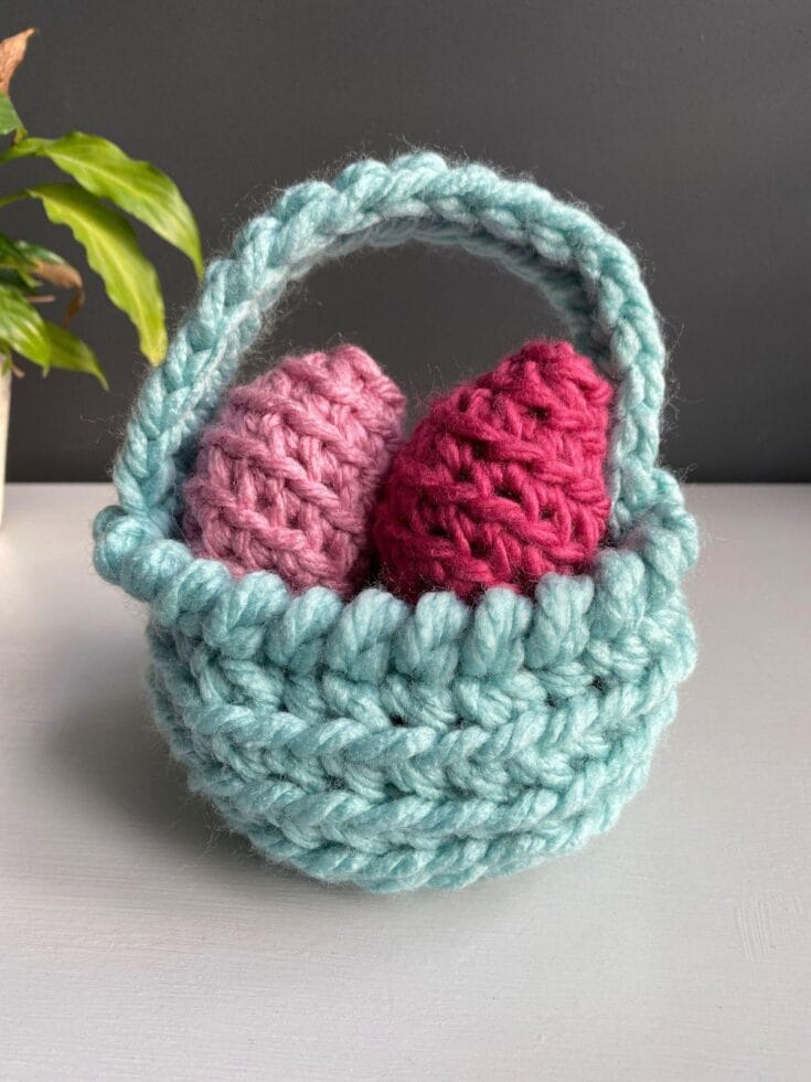 A green Easter Basket made of yarn has 2 pink crochet eggs inside and sits on a white surface