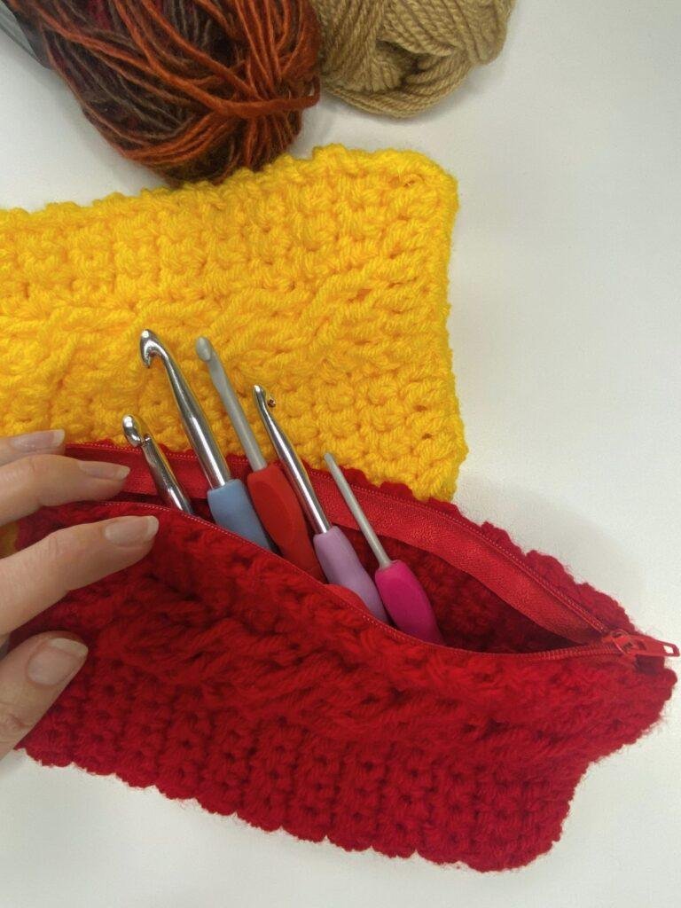 A hand holds a red crocheted case open showing crochet hooks inside. A yellow crochet holder and yarn is behind this