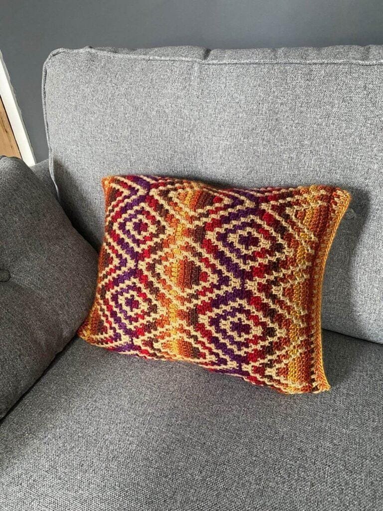 A tribal geometric crochet cushion in bright colours is sat on a grey sofa, with a dark grey wall behind