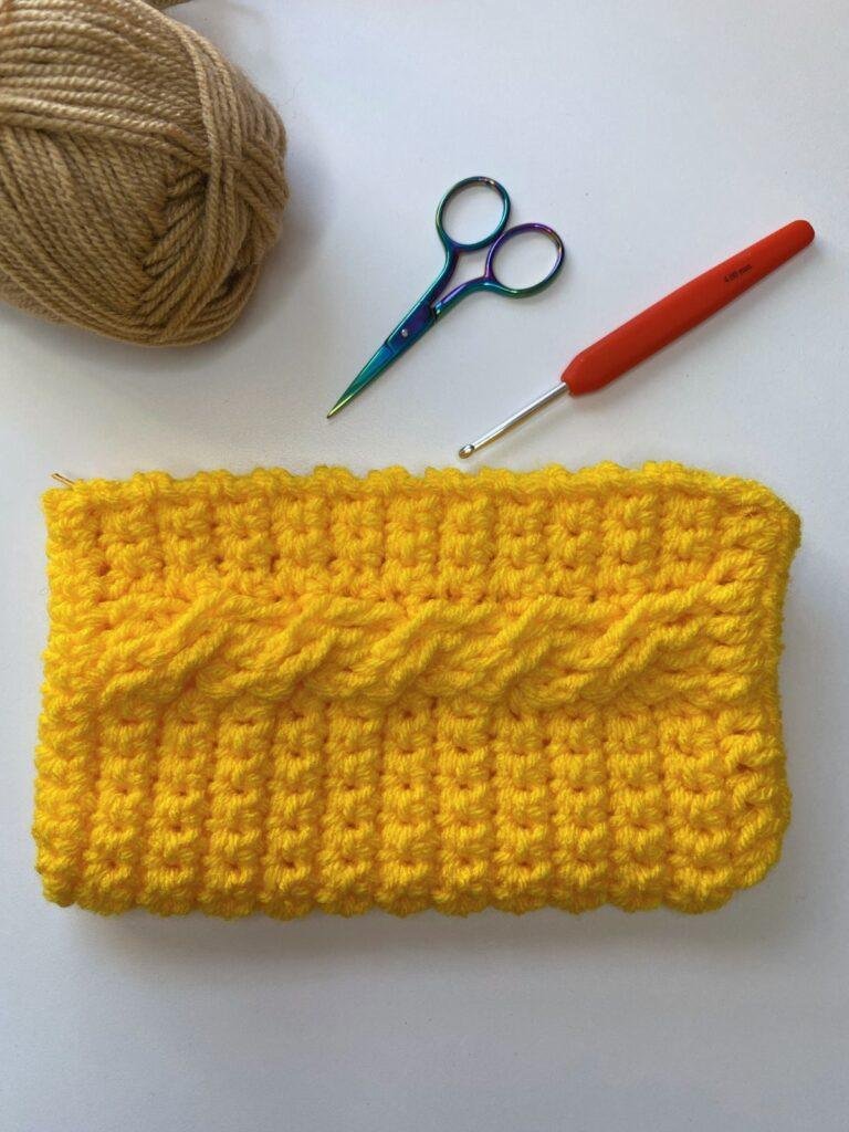 A yellow crochet hook case with cables on the front sits next to scissors, hook and yarn