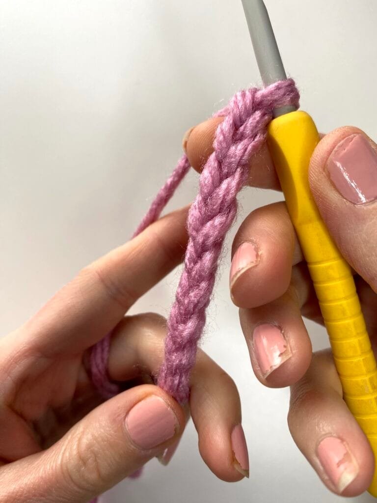 2 hands hold a chain of pink crochet and a yellow crochet hook
