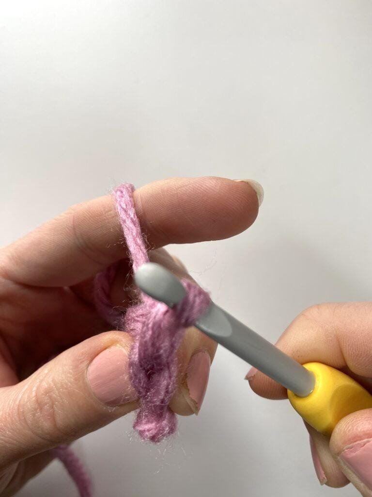 a crocheter chaining 1 after making a bean stitch with a yellow hook and pink yarn