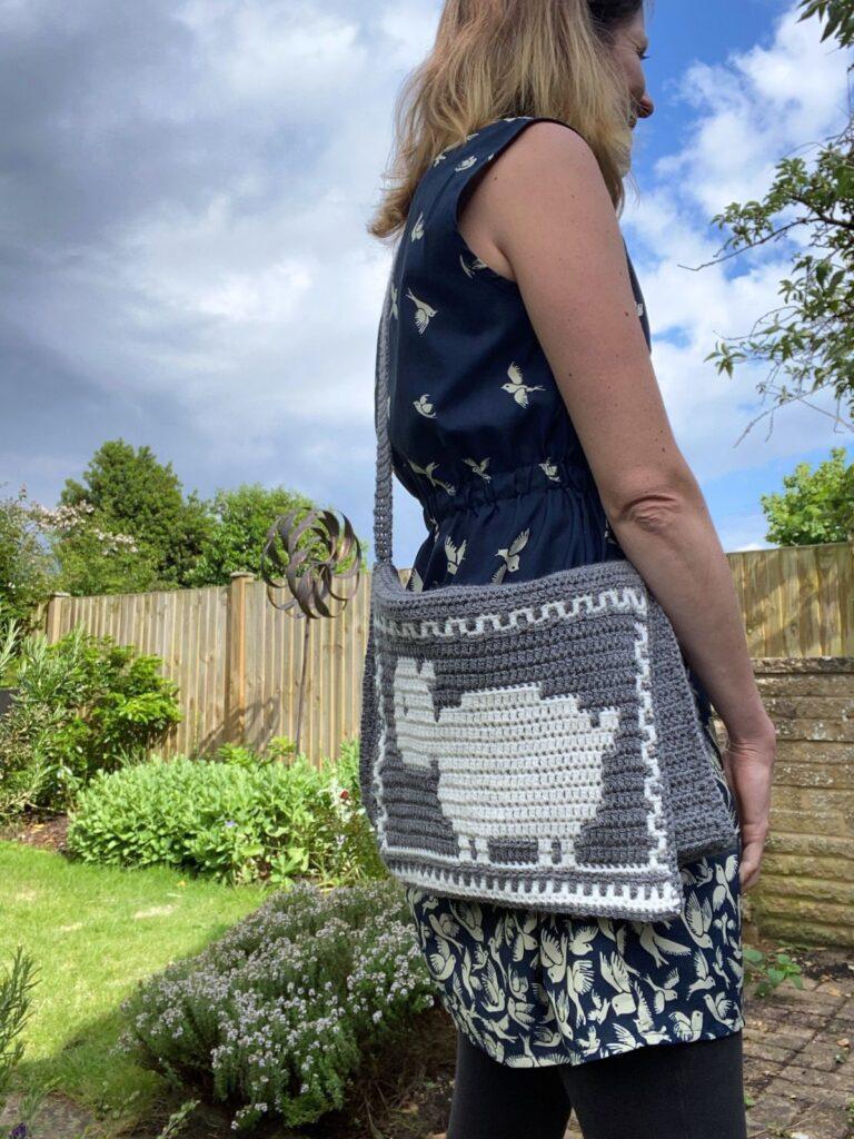 A person stands outside in a garden in a dress with a grey and white crochet sheep bag across their shoulder