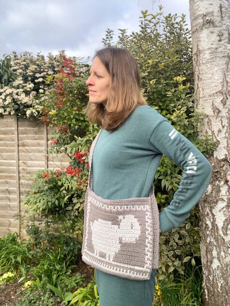 A woman stands in a garden with her hands on her hip wearing a crochet grey shoulder bag with a sheep on it