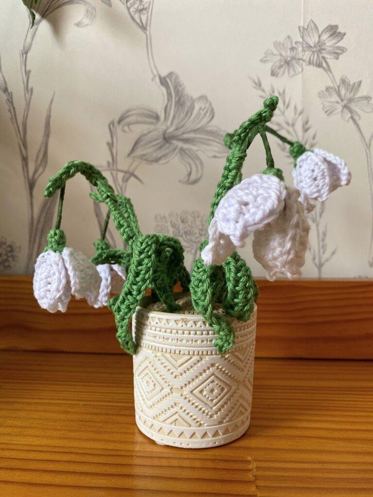 A cream pot holds a set of crochet snowdrops, sat on a wooden shelf against a flowery wallpaper