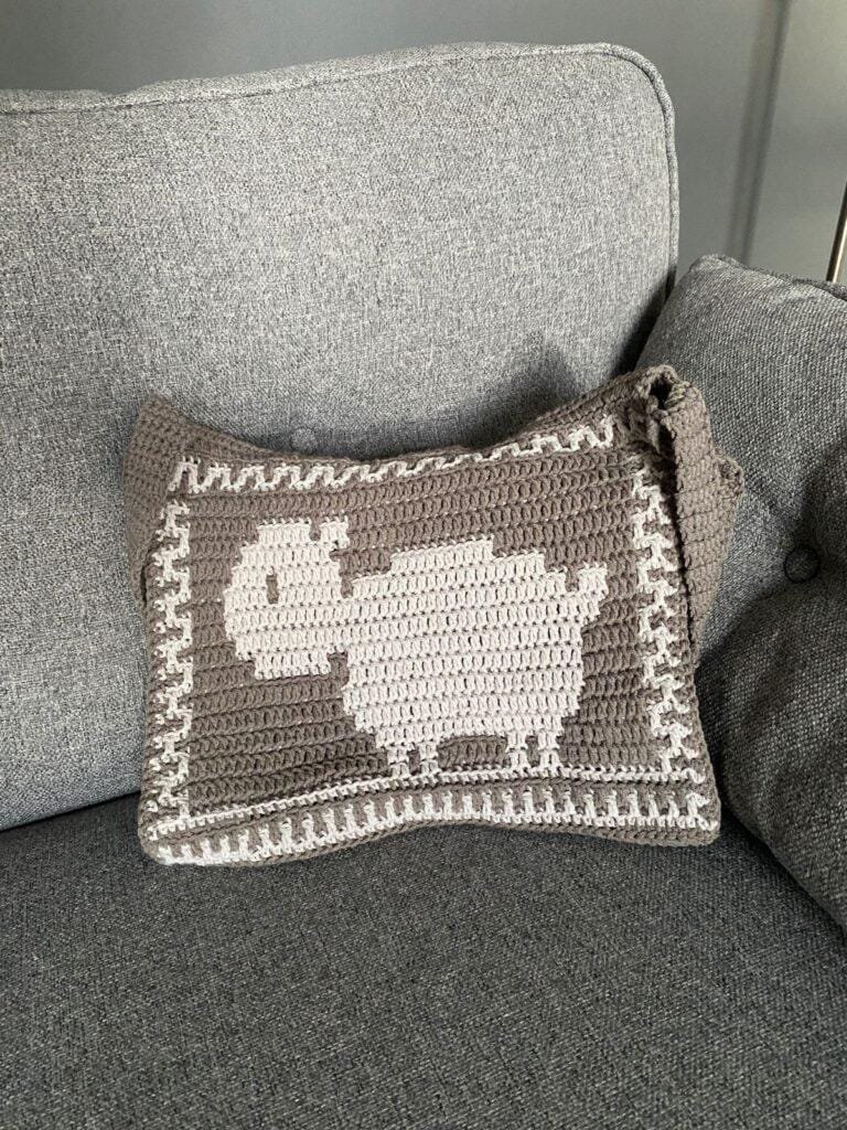 A sheep crocheted onto a grey bag is sat on a grey sofa